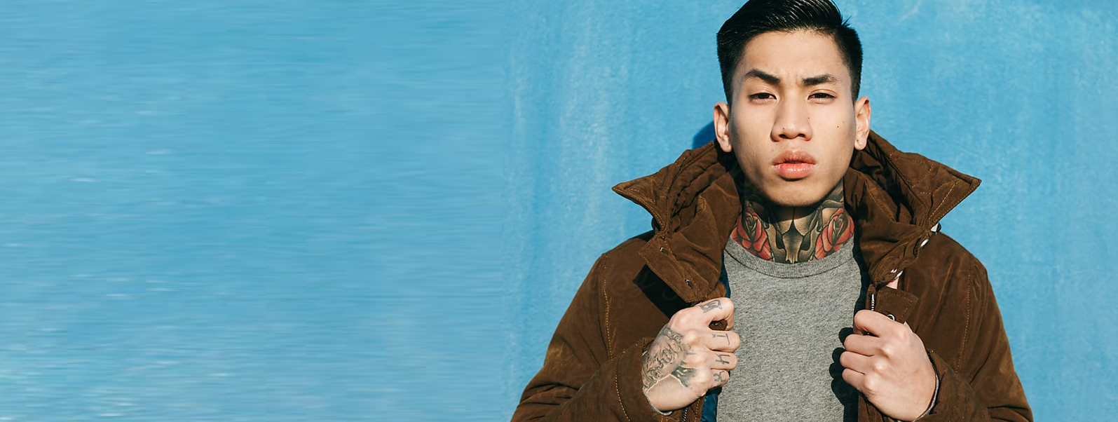 An asian man wearing a brown coat stands in front of a blue wall with tattoos on his hand and neck.