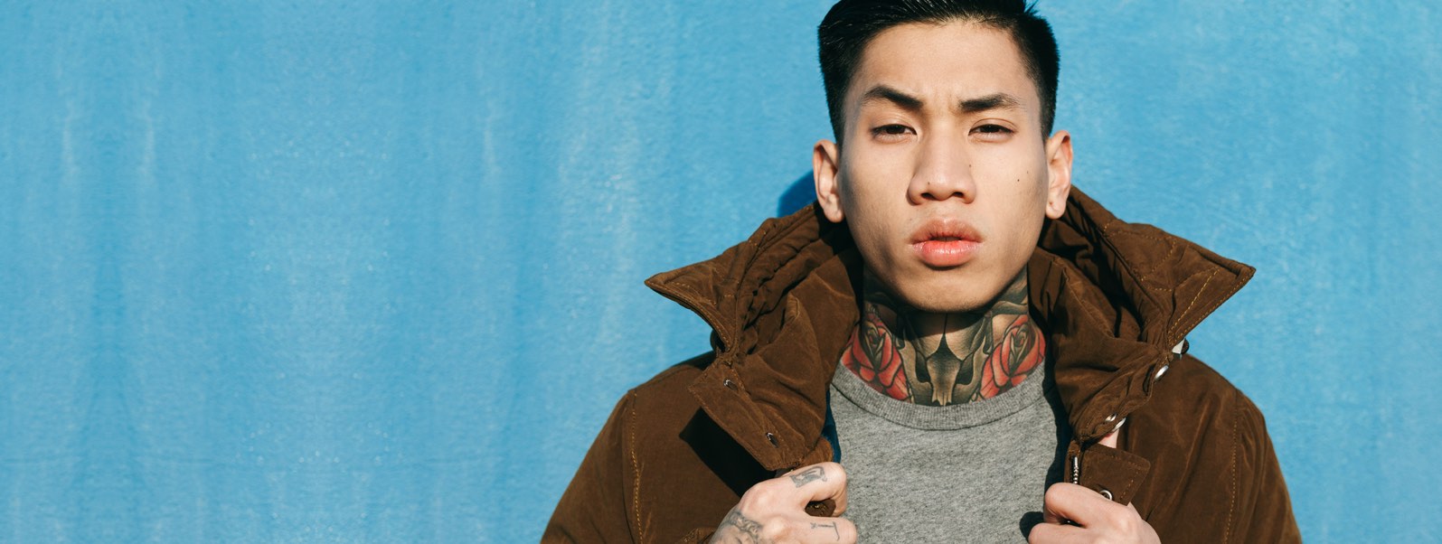 An asian man wearing a brown coat stands in front of a blue wall with tattoos on his hand and neck.