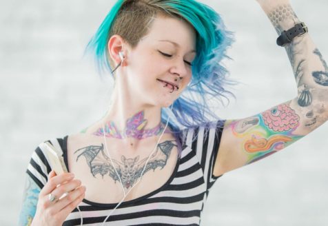 A woman with many facial piercing and tattoos listens to music through ear buds.